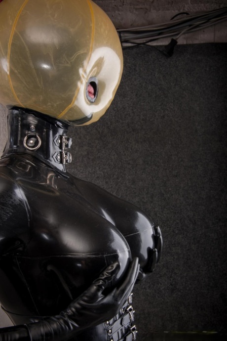 Latex attired slave is affixed with an inflatable hood in dungeon setting
