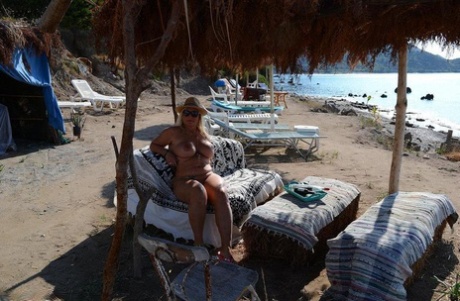 Thick blonde woman with big boobs wanders around a nude beach in a straw hat