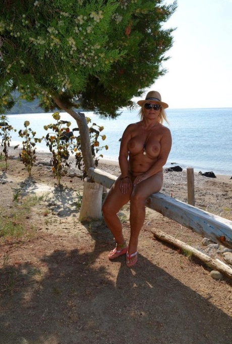 Thick blonde woman with big boobs wanders around a nude beach in a straw hat